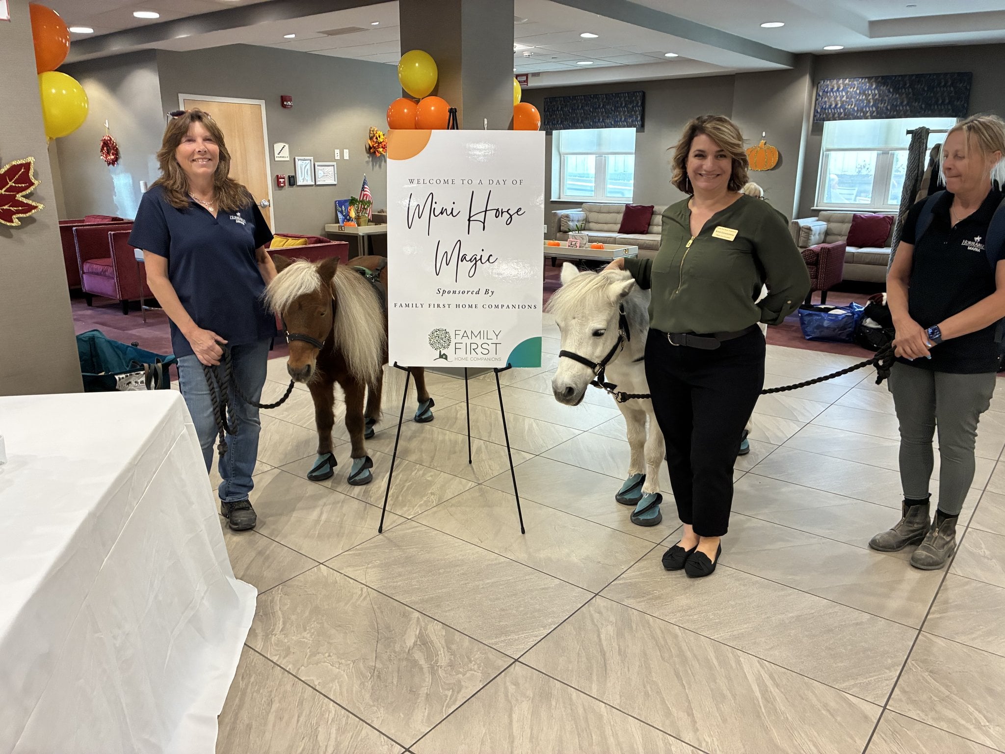Family First Home Companions team with two miniature horses beside the Mini Horse Magic welcome sign at The Five Towns Premier